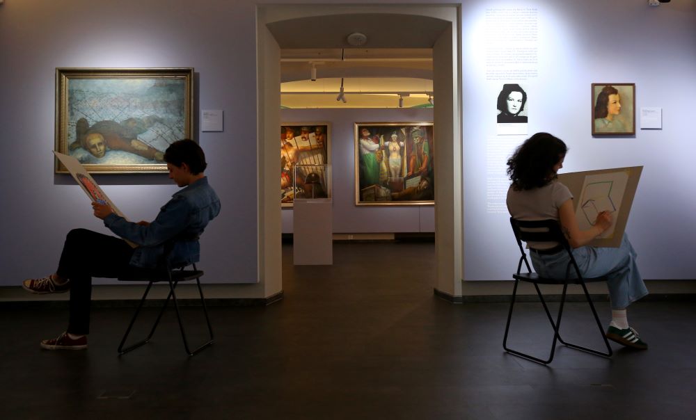 Dve osebi na razstavi Kruha in iger sedita sredi prostora in rišeta.