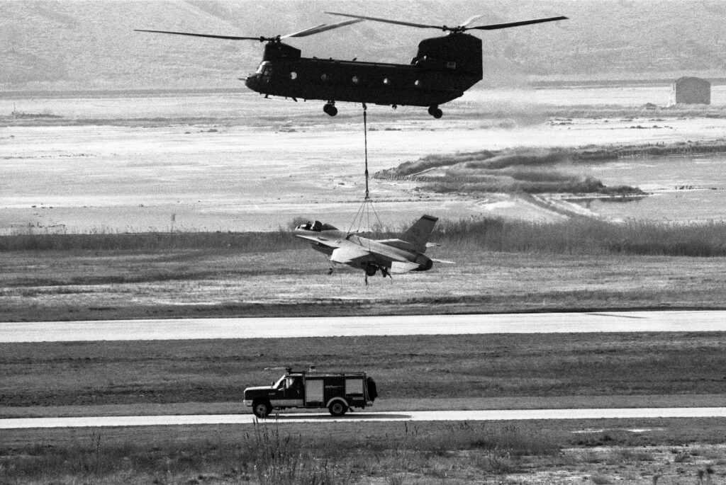 Helikopter z dvema propelerjema leti z navezanim vojaškim letalom, pod njima vozi gasilsko vozilo.