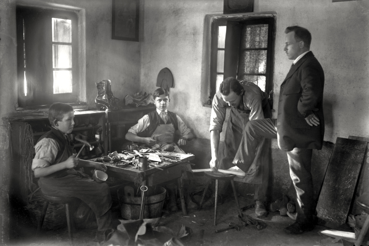 Matevževa delavnica (soba). Fotografija prikazuje čevljarsko delavnico v kateri čevljar ravno meri nogo za izdelavo čevlja. Dva mlajša dečka pa izdelujeta vsak svoj čevelj. Vidni svetlobni žarki, ki prodirajo skozi okno.