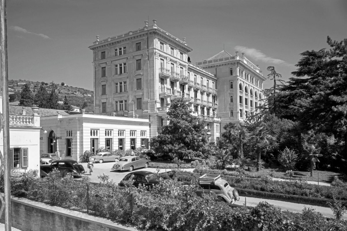 Pogled na poslopje Hotela Palace. Pred hotelom so parkirani različni stari avtomobila. Na desni strani fotografij je viden urejen vrt s palmami.