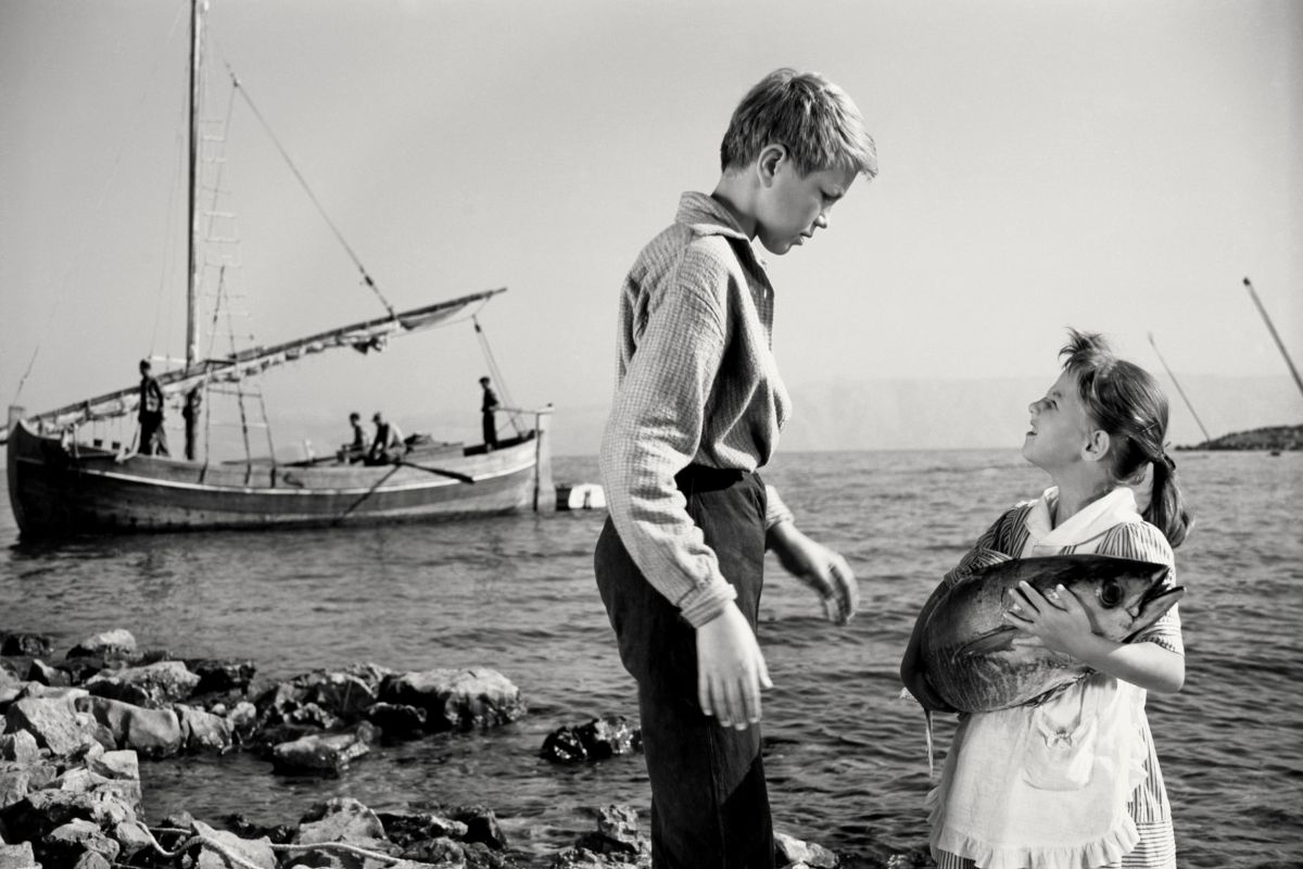 Igralca Tomaž Pesek in Eveline Wohlfeiler v filmu Dobro morje. Deklica v roki trži tuno. Zadaj na fotografiji viden ribiški čoln s posadko.