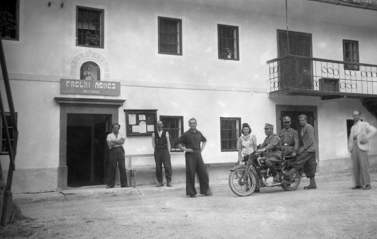 Fotografija ljudi v civilu in uniformah pred gostiščem. Eden izmed moških v uniformi sedi na motorju.