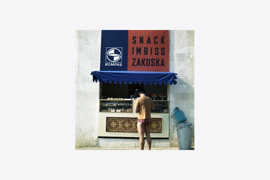 Na fotografiji je stojnica podjetja Kompas, nad njo je na desni napis v treh jezikih »Snack, Imbiss, Zakuska«, levo pa znak- logotip Kompas. Prodajajo se različni sladki prigrizki. Spredaj stoji kupec v majici s kratkimi rokavi in kopalkah.