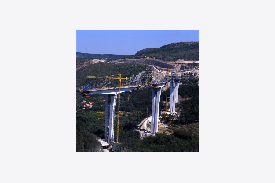 Na voziščni konstrukciji so postavljeni vsi visoki betonski stebri, ob njih visoki žerjavi, v ozadju pa je viden pripravljen teren za nadaljevanje avtocestnega odseka po koncu viadukta.